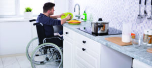 Handicapped Man Sitting On Wheelchair Washing And Cleaning Dishes In Kitchen